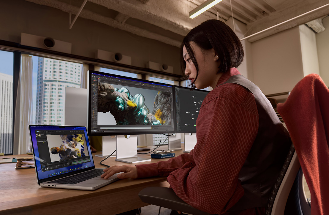 Una persona trabaja en un escritorio con un MacBook Pro conectado a un monitor externo, ambas pantallas muestran un renderizado 3D, se ven edificios a través de la ventana de atrás