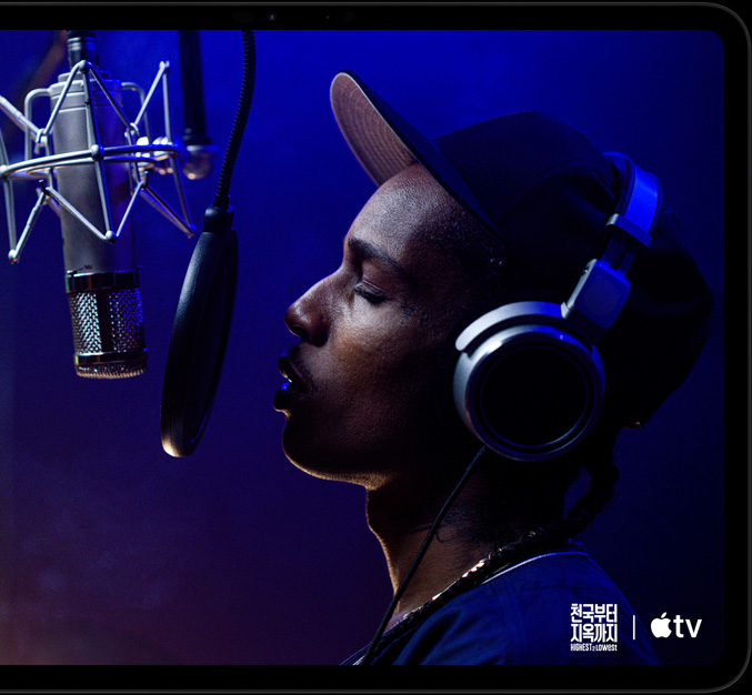 가로 방향으로 놓인 스페이스 블랙 색상 iPad Pro의 앞면. 둥근 형태의 모서리, 블랙 디스플레이 베젤이 보이고 화면에는 Apple TV 오리지널 영화 '천국부터 지옥까지' - Highest 2 Lowest에 등장하는 Yung Felon 역의 에이셉 라키가 마이크 앞에서 랩을 하고 있는 모습이 재생되고 있습니다.