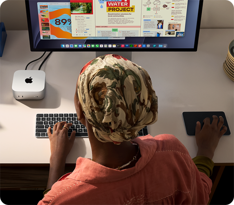Persona pie rakstāmgalda raksta ar Mac mini, kas savienots ar Studio Display, Magic Keyboard tastatūru un Magic Trackpad skārienpaliktni, apkārt dažādi priekšmeti