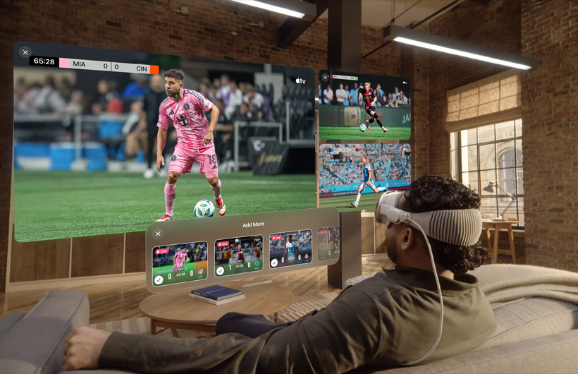 A person wearing Apple Vision Pro is sitting on a couch in a living room, displayed in front of them is a large video screen alongside two smaller screens, each showing a different sporting event. Below the large screen is a selection window that reads: Add More