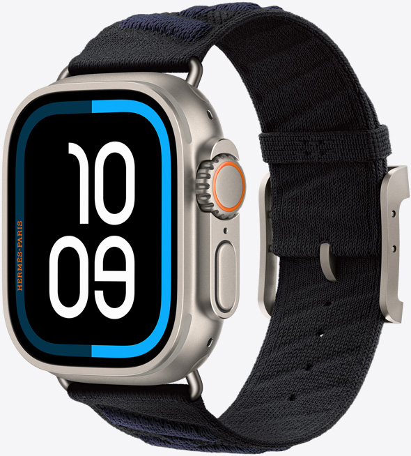 Apple Watch Hermès Ultra 3, titanium case, natural color, edge-to-edge display, rounded corners, right side exterior, Digital Crown, microphone, raised side button, Maritime digital watch face (10 09 digits), paired with En Mer band, noir/bleu nuit color (black with dark blue diagonal textured stripes), knit polyamide yarn material, adjustment holes centered, natural color titanium buckle and hardware