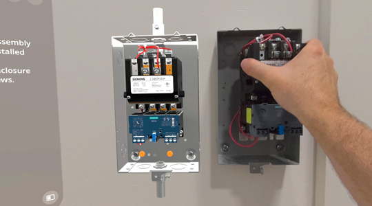 A hand installs a mechanical box with wires onto a wall, near a 3D projection of the same device