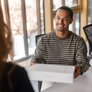 A smiling person in a striped shirt receiving a MacBook Air box in a bright indoor setting.