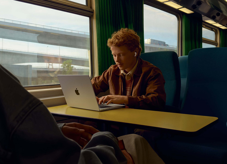 기차의 창가 좌석에 앉아 AirPod을 착용한 채 MacBook으로 작업중인 사람의 모습