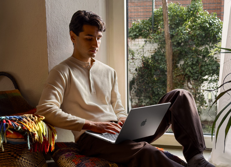 창문 옆에서 바닥에 앉아 MacBook을 사용하는 사람의 모습.