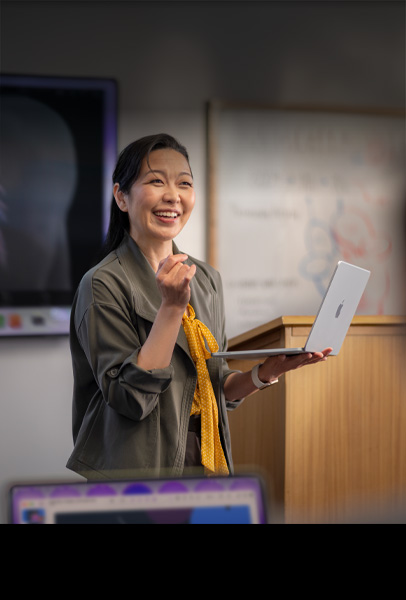 A professor gives a presentation to their students in a lecture hall while holding a silver MacBook, demonstrating performance and long battery life