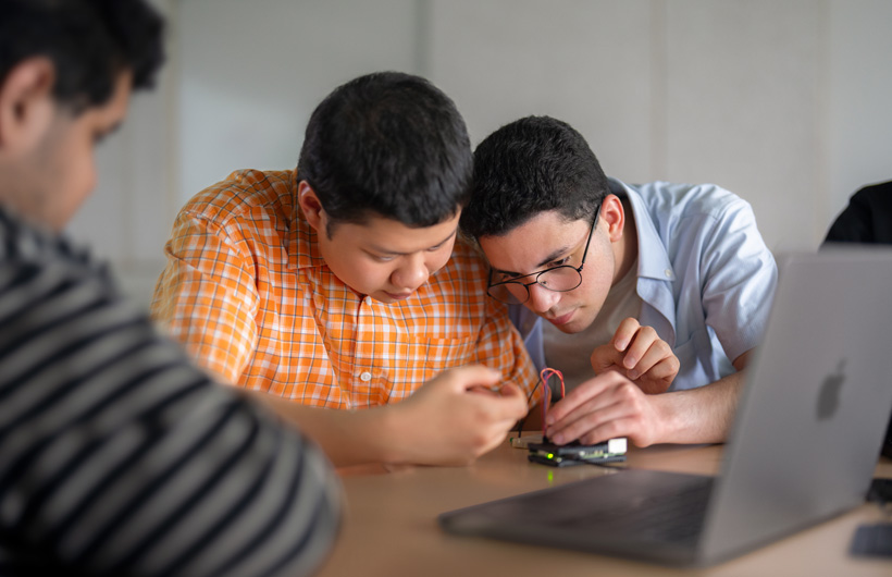 Deux personnes scrutent une carte de circuit imprimé à côté d’un MacBook, tandis qu’une troisième les observe.