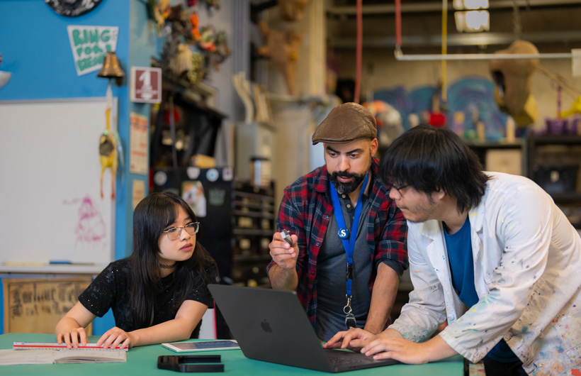 Trois personnes sont réunies autour d’un MacBook posé sur une table dans un atelier coloré rempli d’outils et d’œuvres d’art.