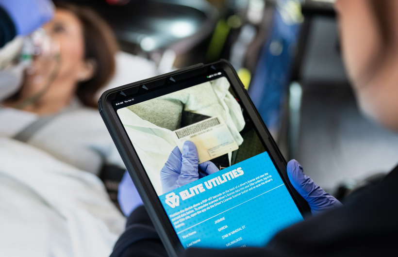 Die Elite Utilities App ist auf dem iPad geöffnet, und ein Sanitäter scannt den Führerschein einer Patientin, die auf einem Transportbett liegt