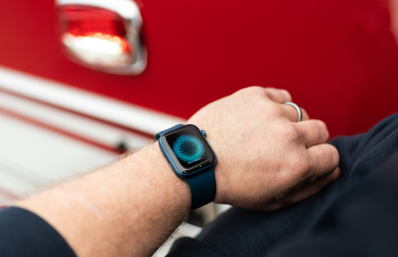 Eine Apple Watch an einem Handgelenk zeigt die Meldung "Atme ein…" an mit einem Feuerwehrauto in der Nähe