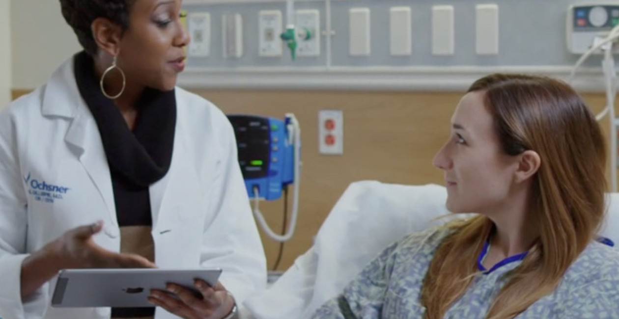 Una persona de un equipo sanitario habla con su paciente a pie de cama mientras sujeta un iPad.