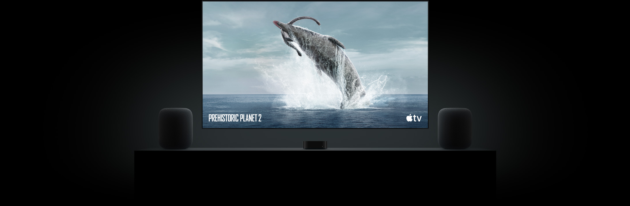 Suuri taulutelevisio, jolla näkyy vaikuttava kuva dinosauruksesta Prehistoric Planet ‑sarjasta. Televisio on Apple TV:n yläpuolella, ja sitä reunustaa kaksi olohuoneen senkin päällä olevaa HomePod-kaiutinta