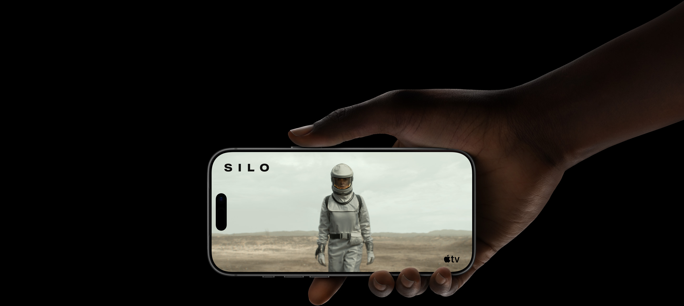 A hand holding iPhone 17 Pro, displaying a woman wearing a protective suit and helmet, walking through a barren landscape. Apple TV logo, series title: Silo