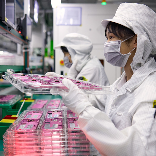 Deux personnes portant des combinaisons de travail couvrant tout le corps manipulent des pièces d’iPhone dans une usine d’assemblage