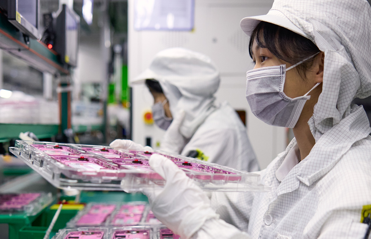 Deux personnes portant des combinaisons de travail couvrant tout le corps manipulent des pièces d’iPhone dans une usine d’assemblage