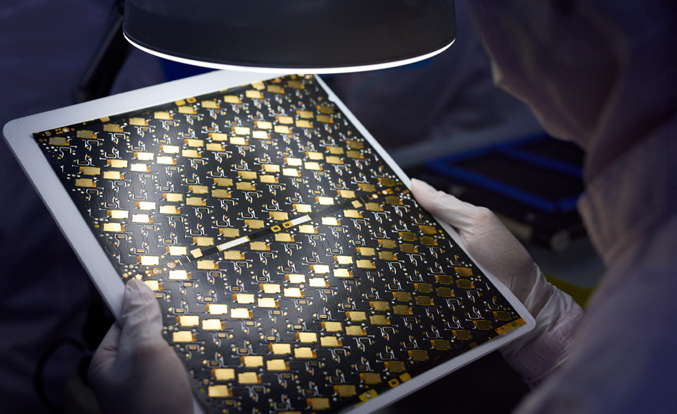 Worker wearing gloves examining a circuit board panel under a magnifying inspection light