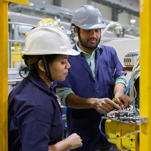 Two employees in work smocks and safety hats stand in front of an assembly machine
